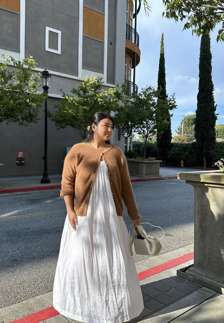 A woman styling a tan cropped knit cardigan over a flowy white maxi dress, holding a cream handbag.