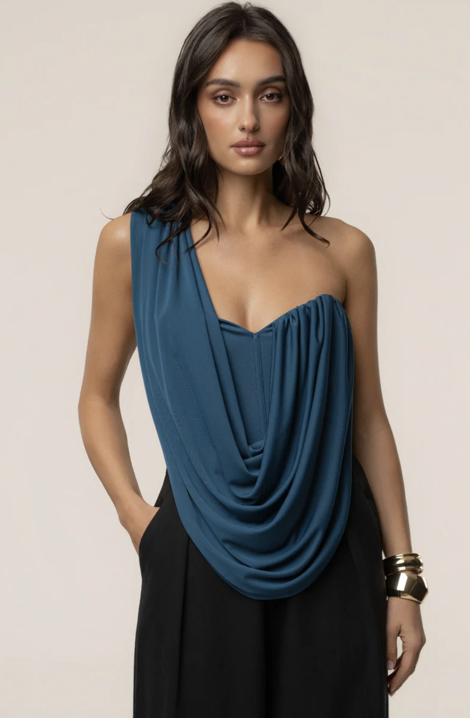 A person wearing a slate blue off-shoulder top with a deep draped cowl neckline, paired with black high-waisted trousers.