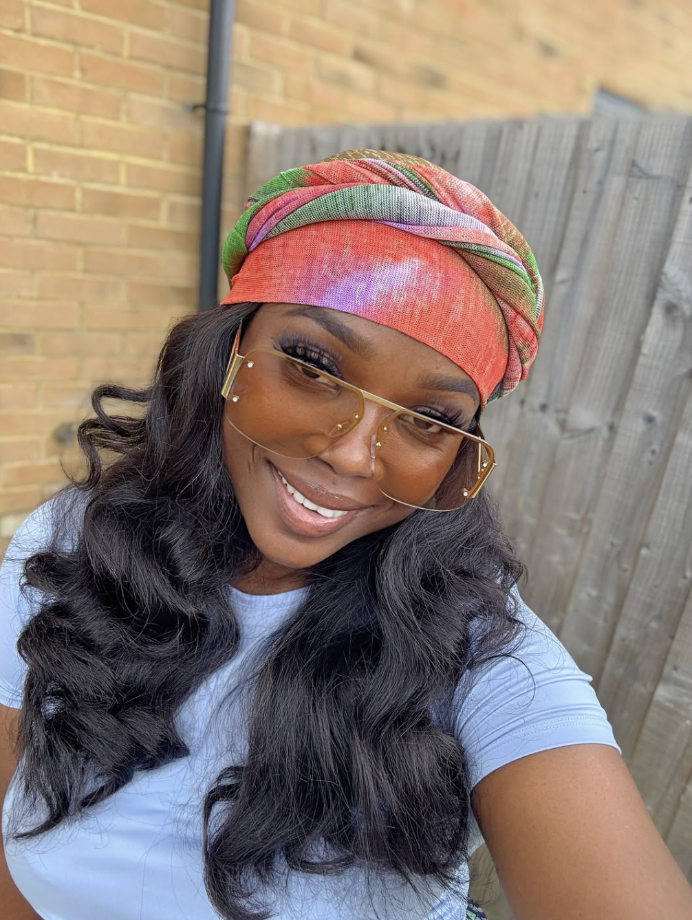 A close-up shot of a smiling woman with long wavy hair, wearing a vibrant pink, green, and orange tie-dye silk scarf styled as a full head wrap, paired with oversized gradient aviator sunglasses.
