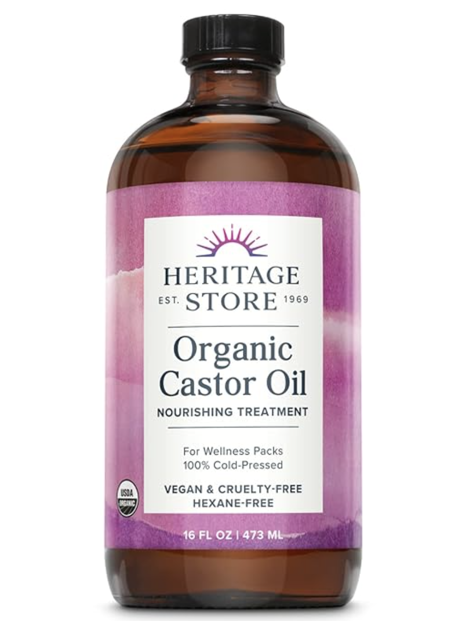 A dark amber glass bottle of Heritage Store Organic Castor Oil with a purple label, highlighting its 100% cold-pressed and hexane-free formula for lash and brow health.