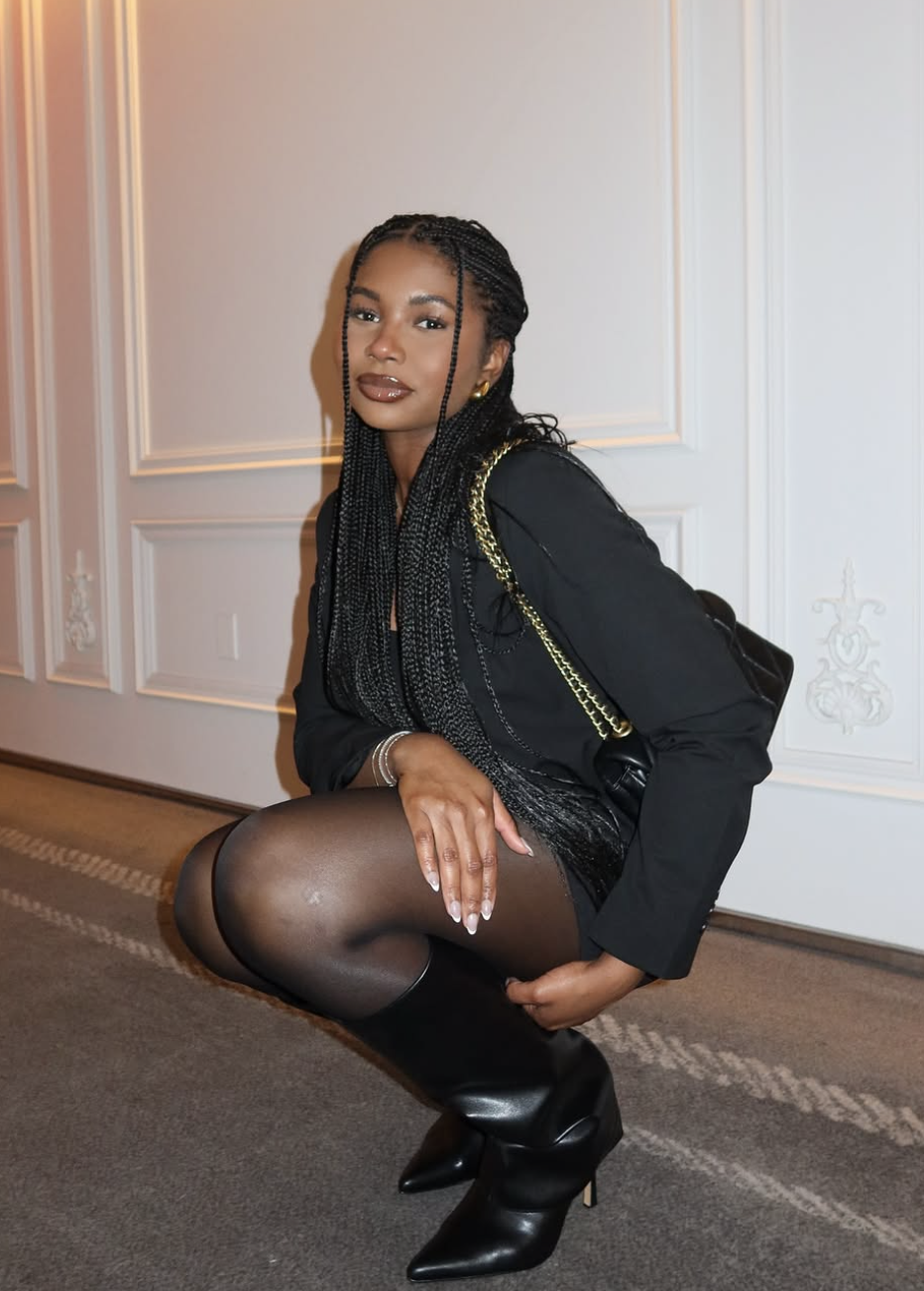 Clarke Peoples with long, small knotless braids, wearing a black blazer and sheer tights, demonstrating how length below the chin balances a heart face shape.