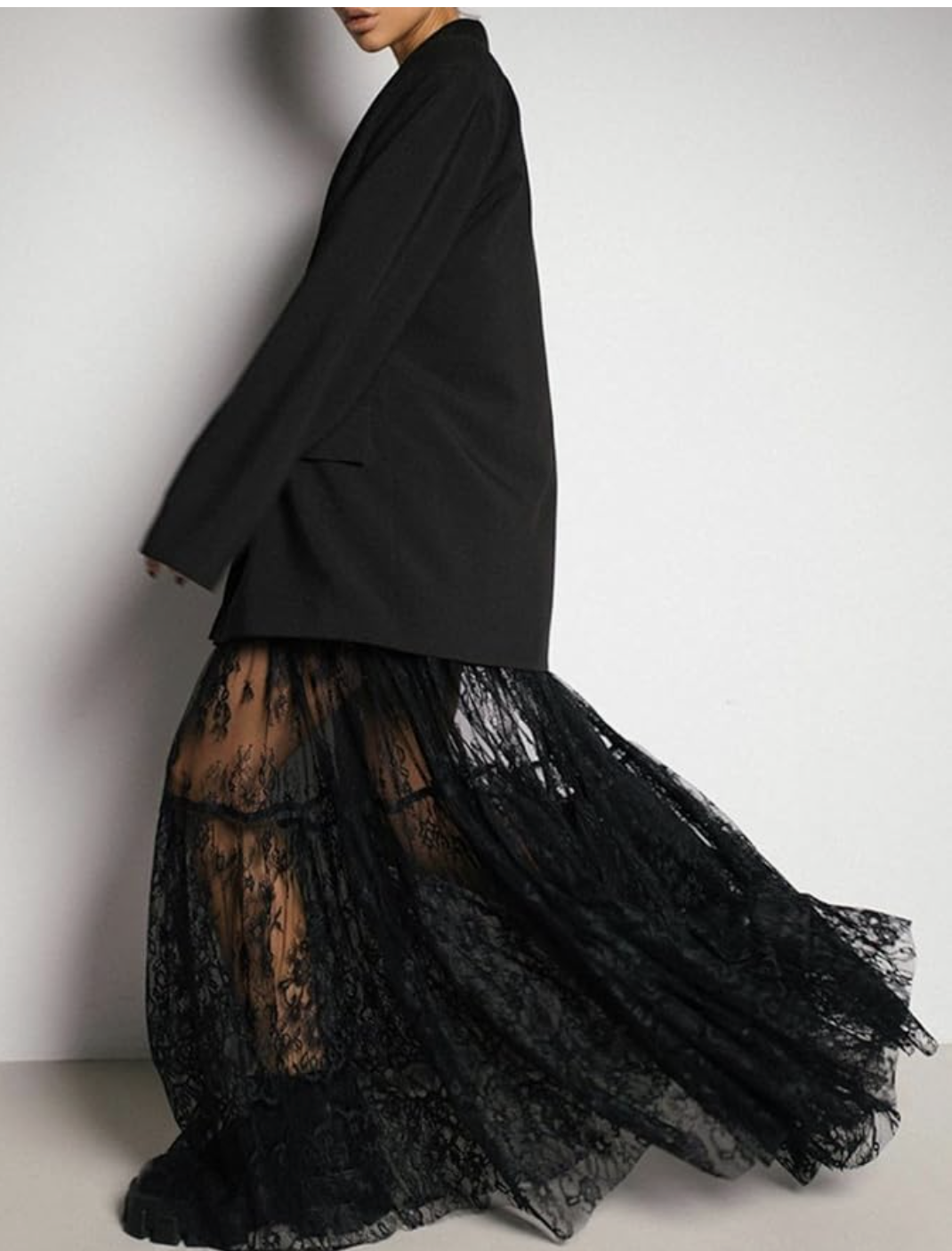 A close-up of a person wearing a sheer black floral lace maxi skirt with a tiered ruffled hem, paired with an oversized black blazer.