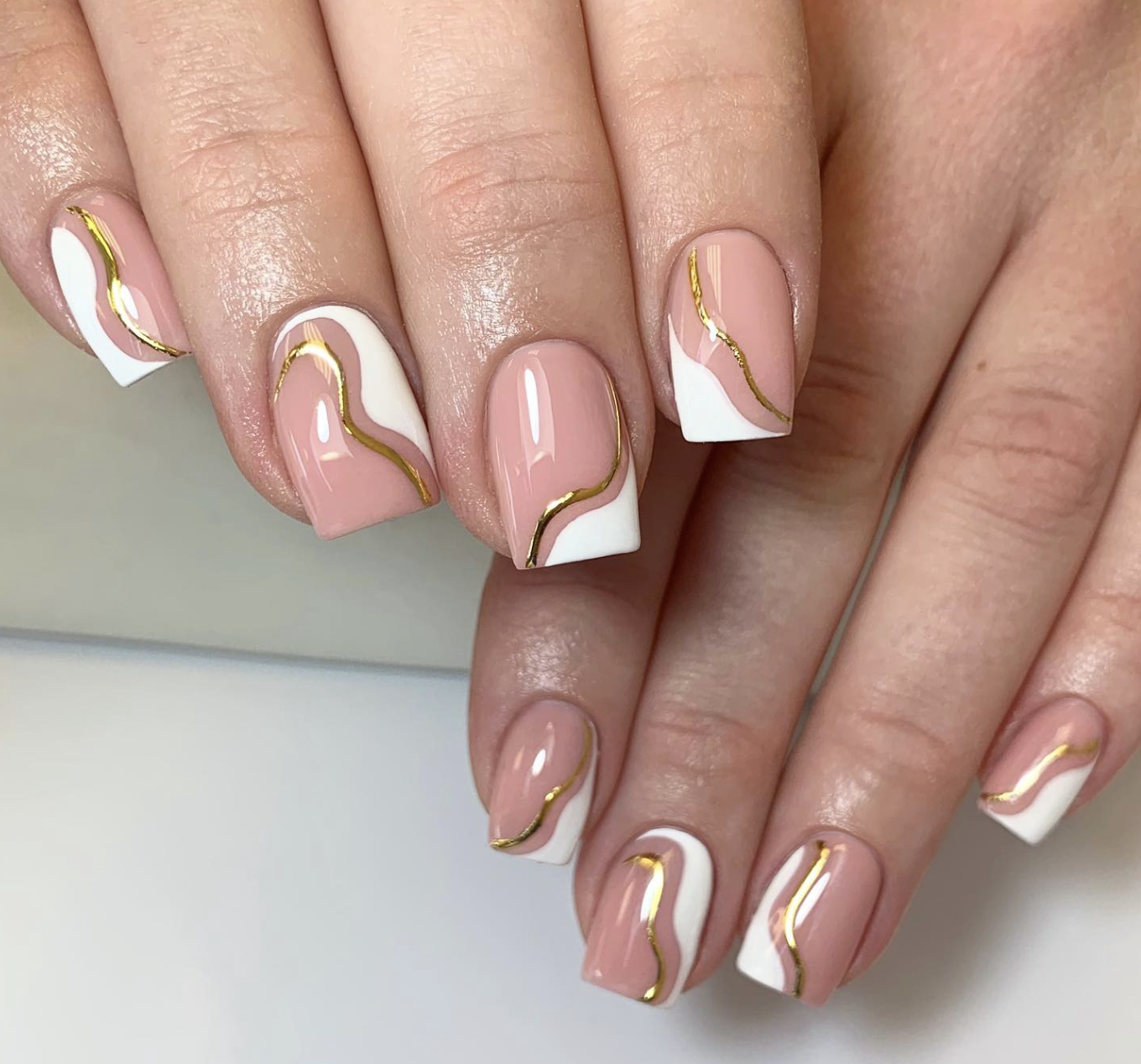 A close-up of a short neutral manicure featuring abstract white swirls and delicate gold foil accents on a nude base.
