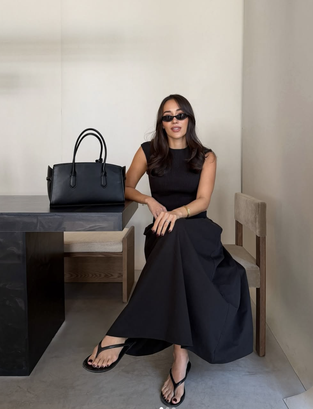 A woman walking in a black midi dress and minimalist black leather flip flops, carrying a woven straw bag.