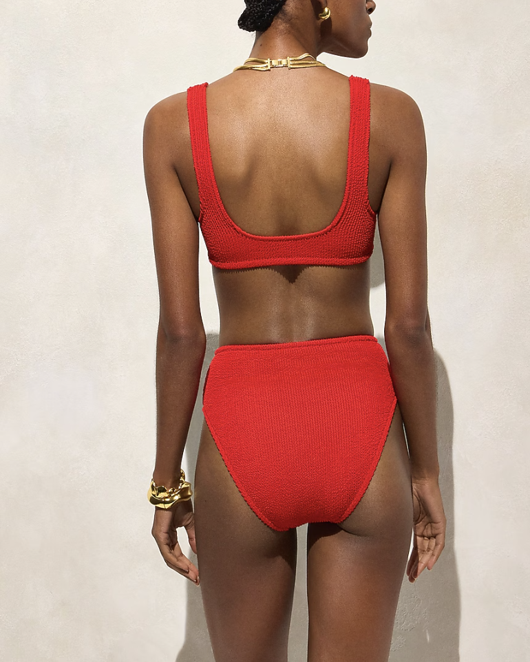 A woman from behind wearing a vibrant red one-piece swimsuit with a low scoop back and textured fabric, accessorized with a chunky gold bracelet.