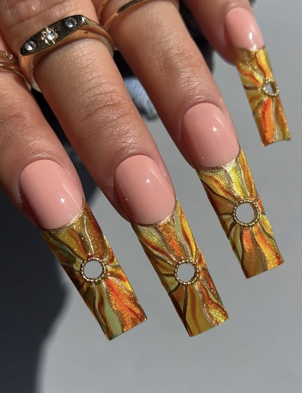 Extra-long stiletto nails with a nude base, featuring large hand-painted sunflower petals in orange and yellow with shimmering gold glitter centers and silver rhinestone outlines.
