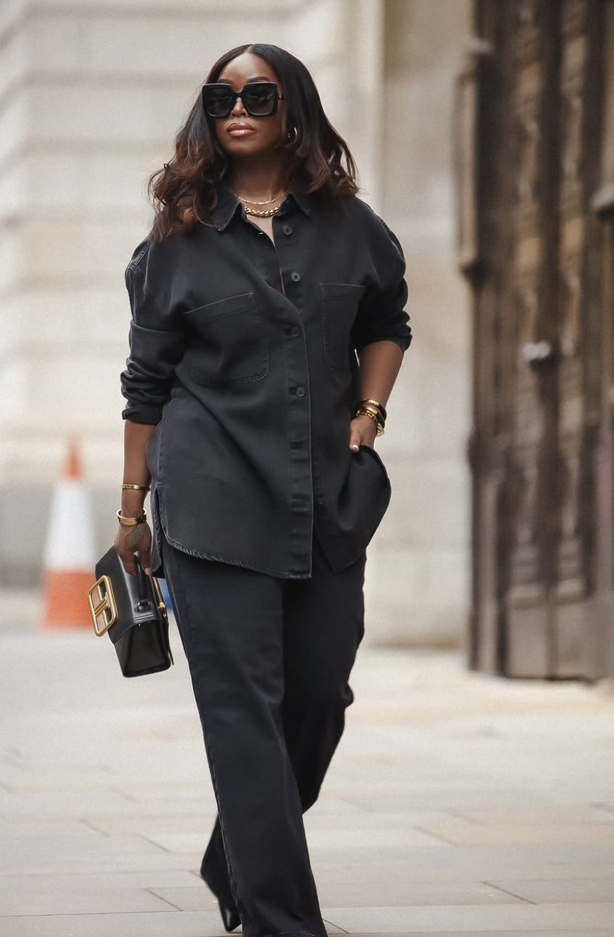 A woman wearing a dark blue denim button-up shirt with black jeans, oversized sunglasses, and carrying a black quilted leather shoulder bag with a gold chain strap.