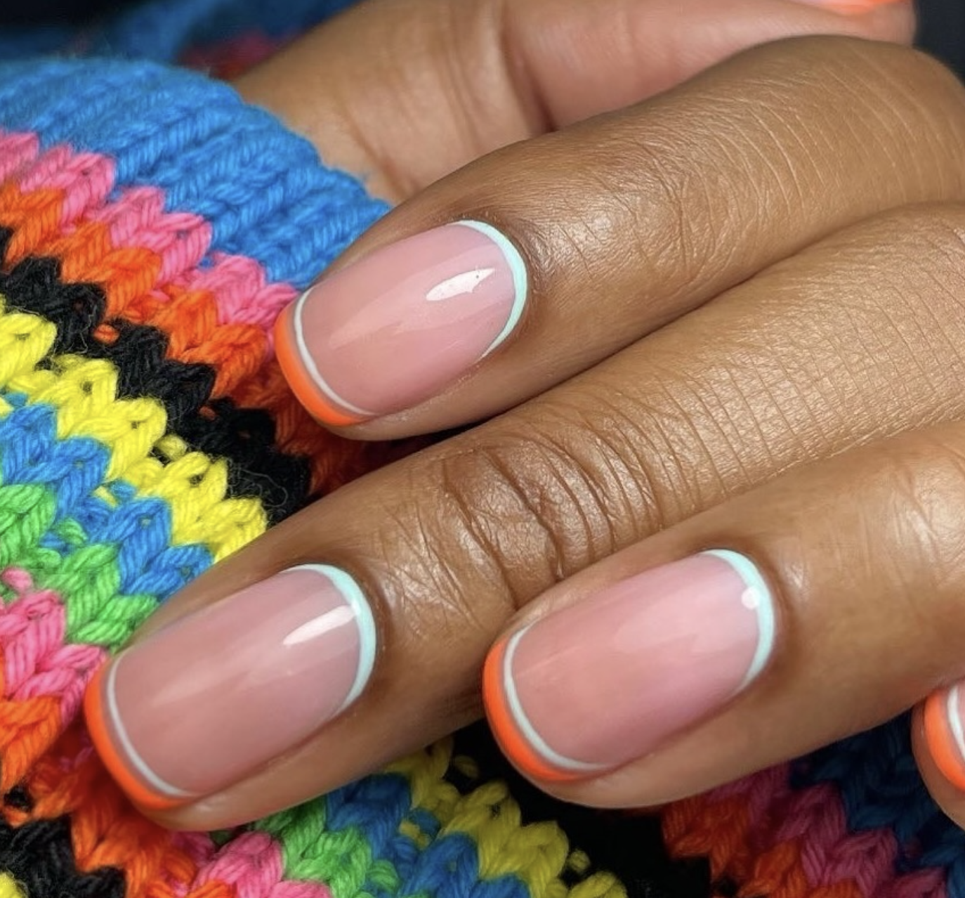 A close-up of short nails featuring a reverse French half-moon design with light blue and neon orange lines around the cuticle bed over a sheer nude base.