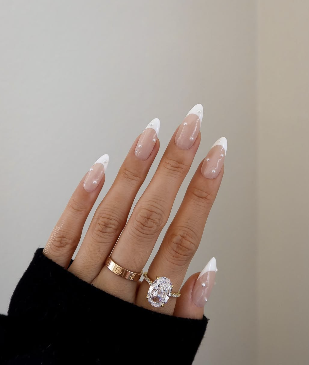 Long almond-shaped nails with white French tips, featuring small white pearl accents placed near the cuticles.