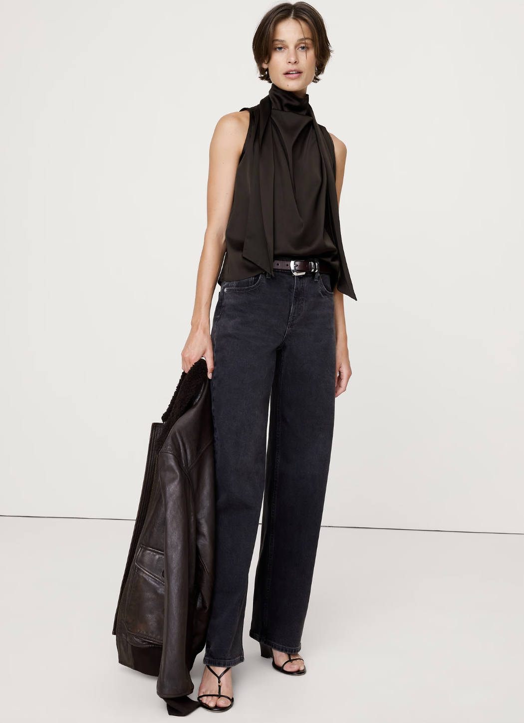 A woman wearing a black sleeveless tie-neck top tucked into dark wide-leg jeans, holding a leather jacket and wearing strappy sandals.