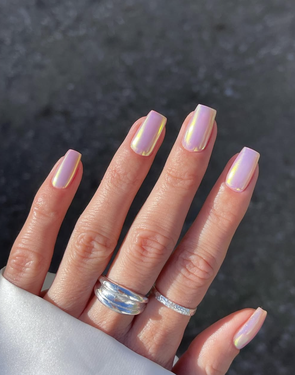A close-up of almond-shaped nails with a light pink base and a holographic chrome finish reflecting pastel rainbow colors.