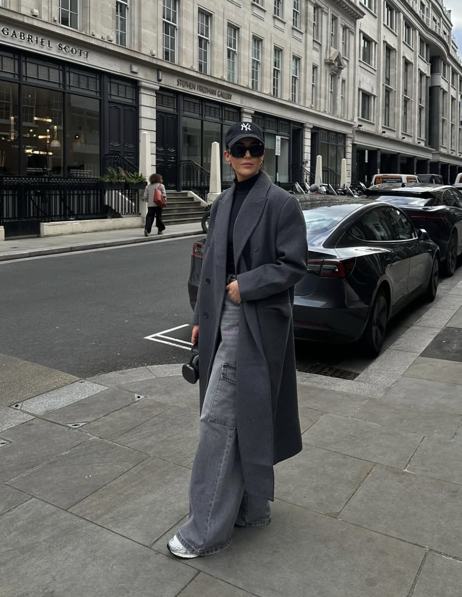 A woman wearing an elevated monochromatic cream pleated outfit featuring a matching neck scarf, a dark chocolate brown suede tote bag, and tortoiseshell sunglasses.