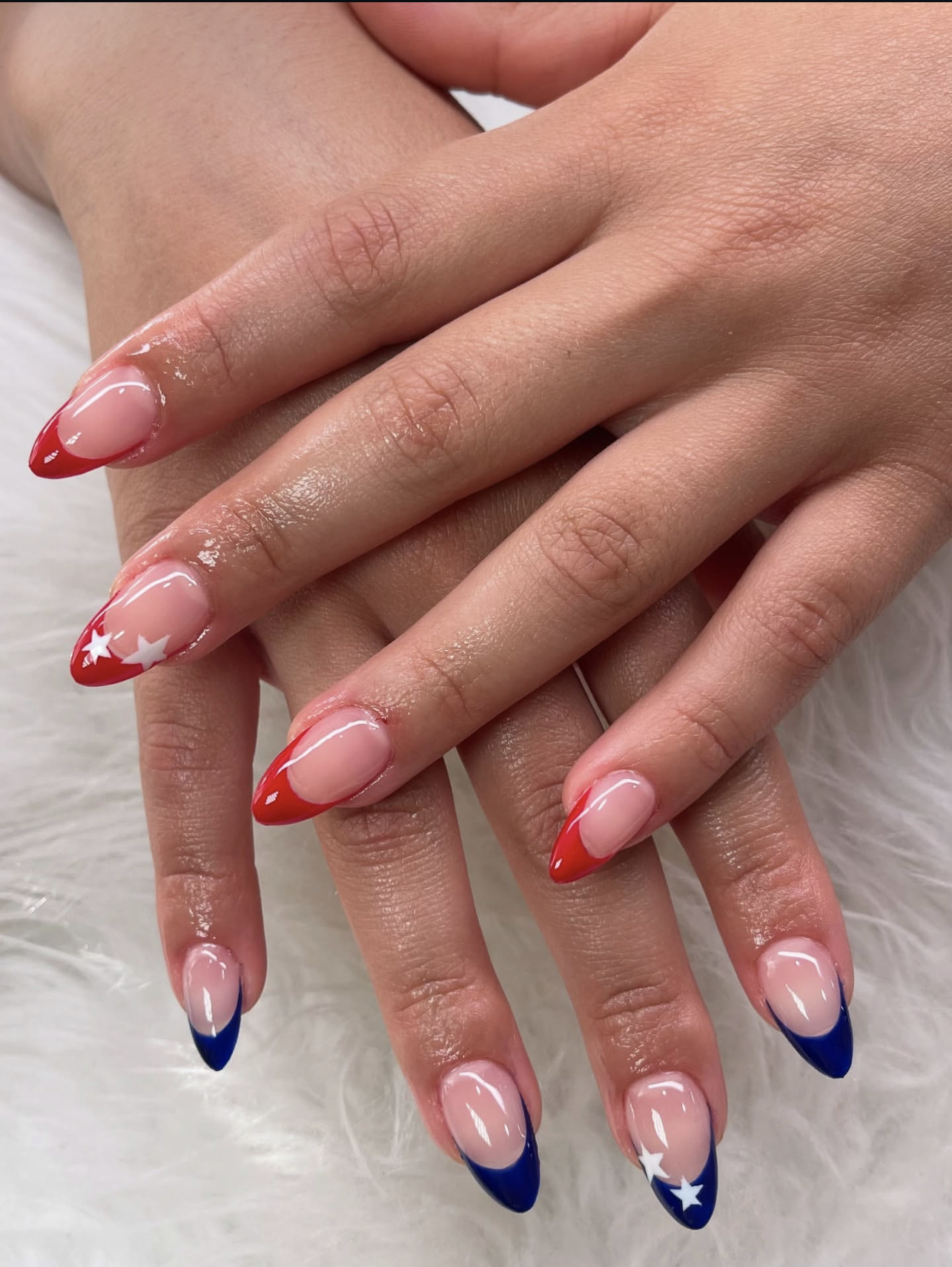 Long, oval-shaped nails featuring a creative 4th of July design with bold red French tips adorned with crisp white stars on one hand, and striking cobalt blue French tips on the other.