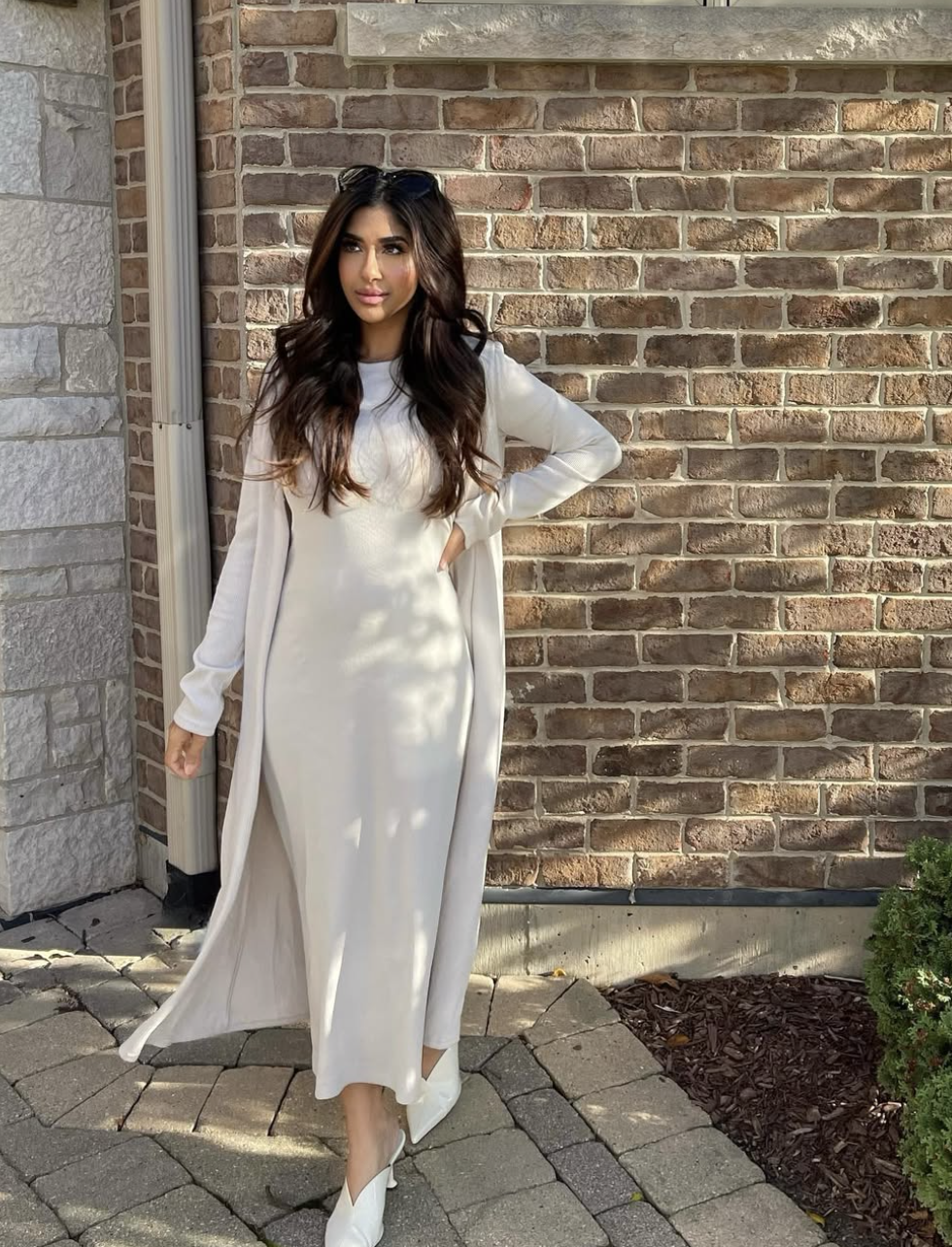 A woman wearing a white long-sleeve bodycon maxi dress layered with a matching floor-length white cardigan, standing against a brick wall.