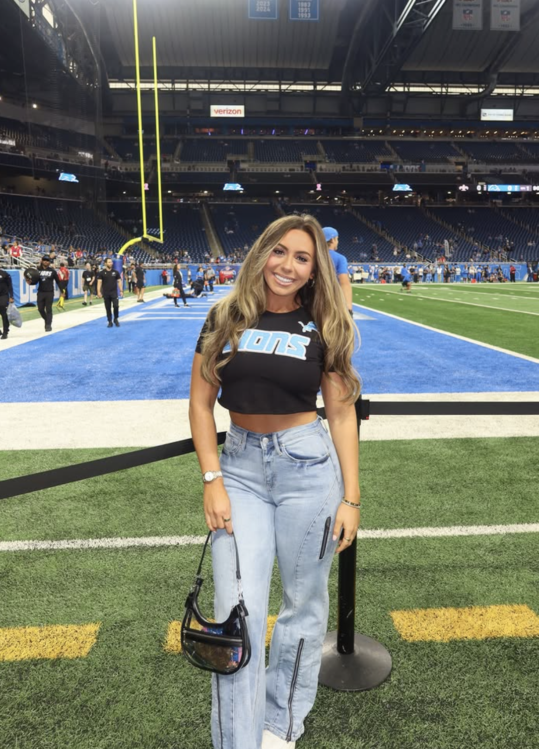 Woman at a Detroit Lions game wearing a black cropped team tee, light-wash utility jeans, white sneakers and a clear stadium bag.