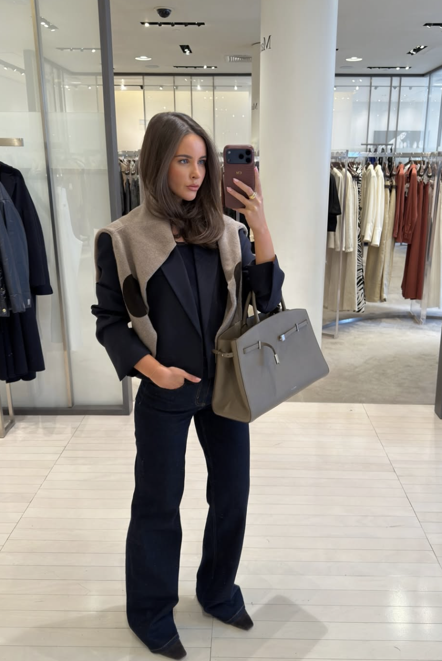 A woman wearing a monochromatic navy blue outfit featuring a structured blazer and trousers, layered with a contrasting beige vest and styled with a grey leather handbag.