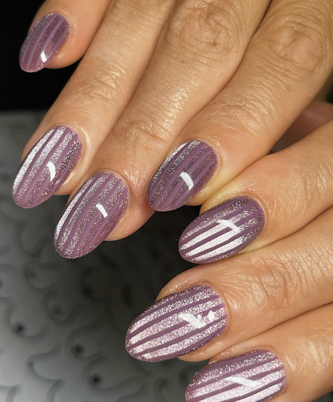 Close-up of striped nail art on oval nails featuring a dusty mauve base with vertical, shimmering silver glitter stripes.