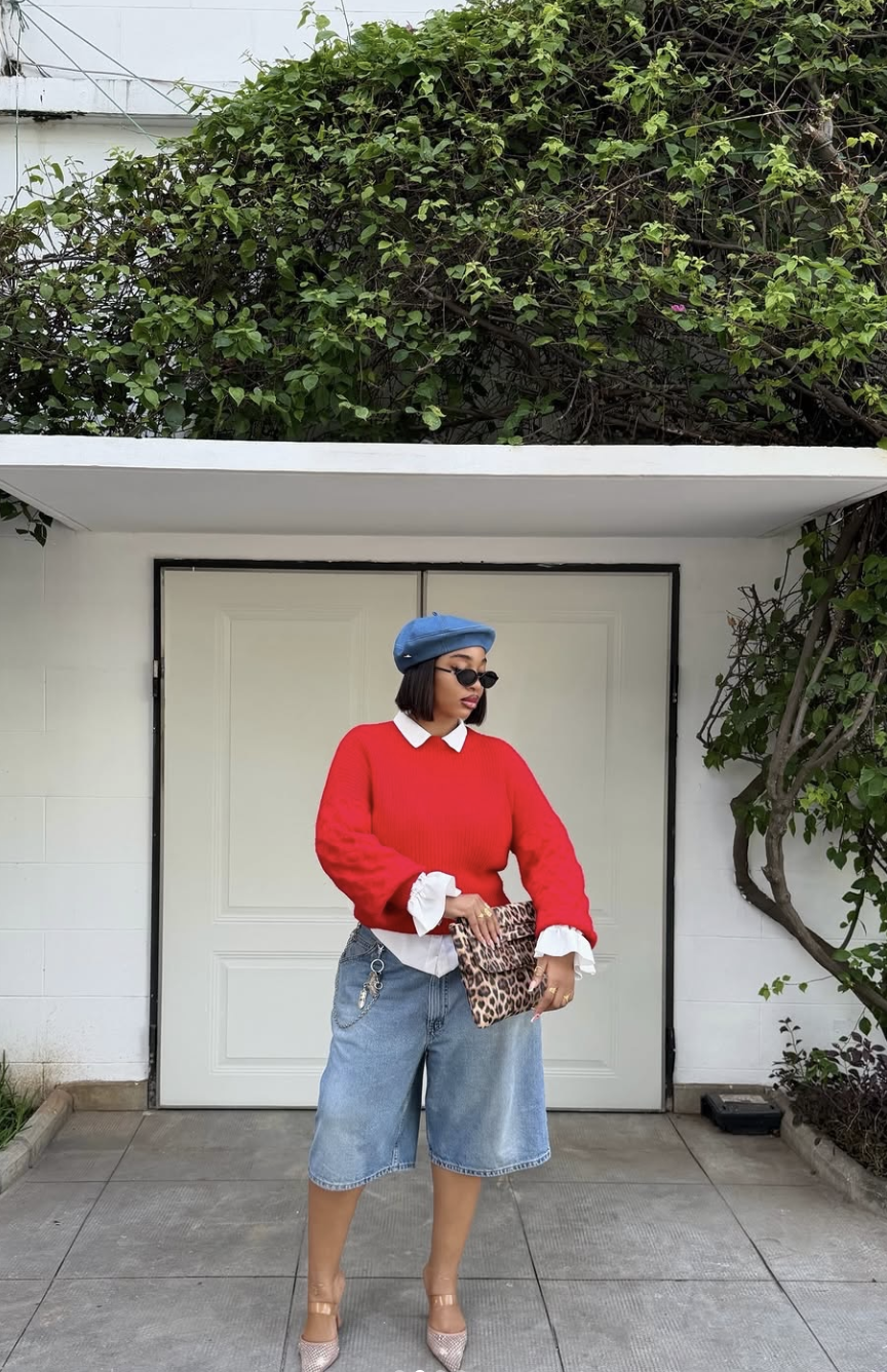 A woman wearing a bright red oversized sweater over a white collared shirt, styled with denim Bermuda shorts, a blue beret, and black pointed-toe mules.