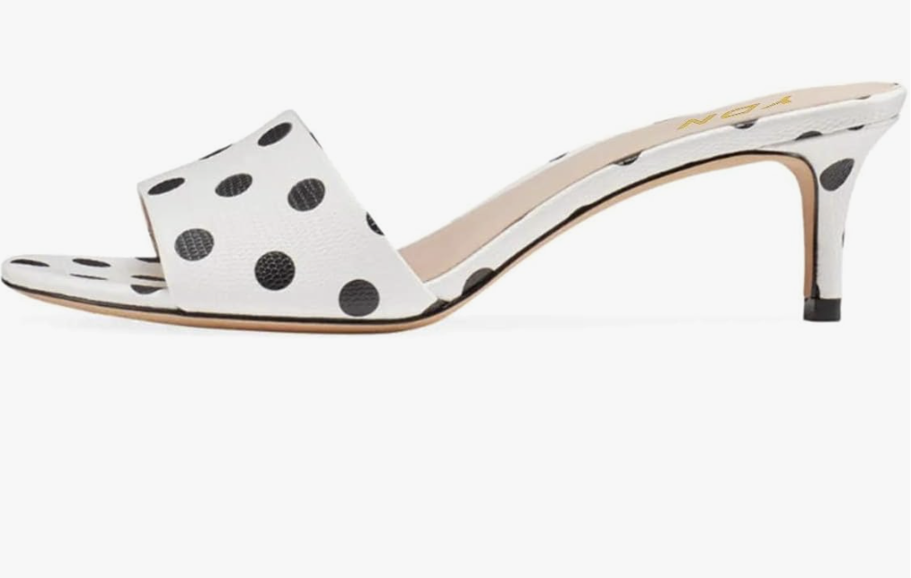 A close-up view of a chic white slip-on mule shoe featuring a low kitten heel and a playful black polka-dot pattern.