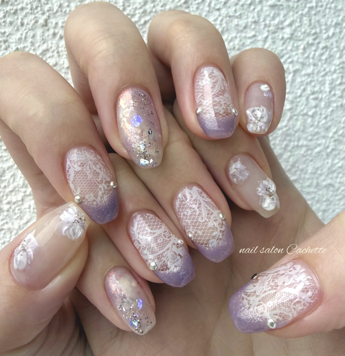 Long almond-shaped lavender nails featuring white lace patterns, iridescent glitter, and small pearl accents.