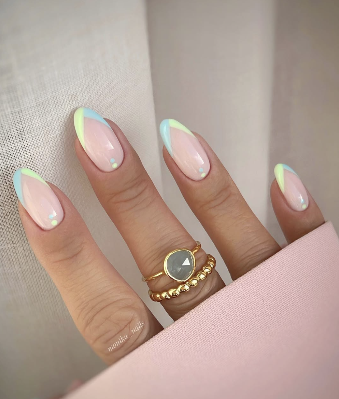 A close-up of a hand with long almond-shaped nails featuring a milky beige base and alternating French tips in shades of aquamarine blue and mint green, accented with tiny holographic sequins and gold rings.