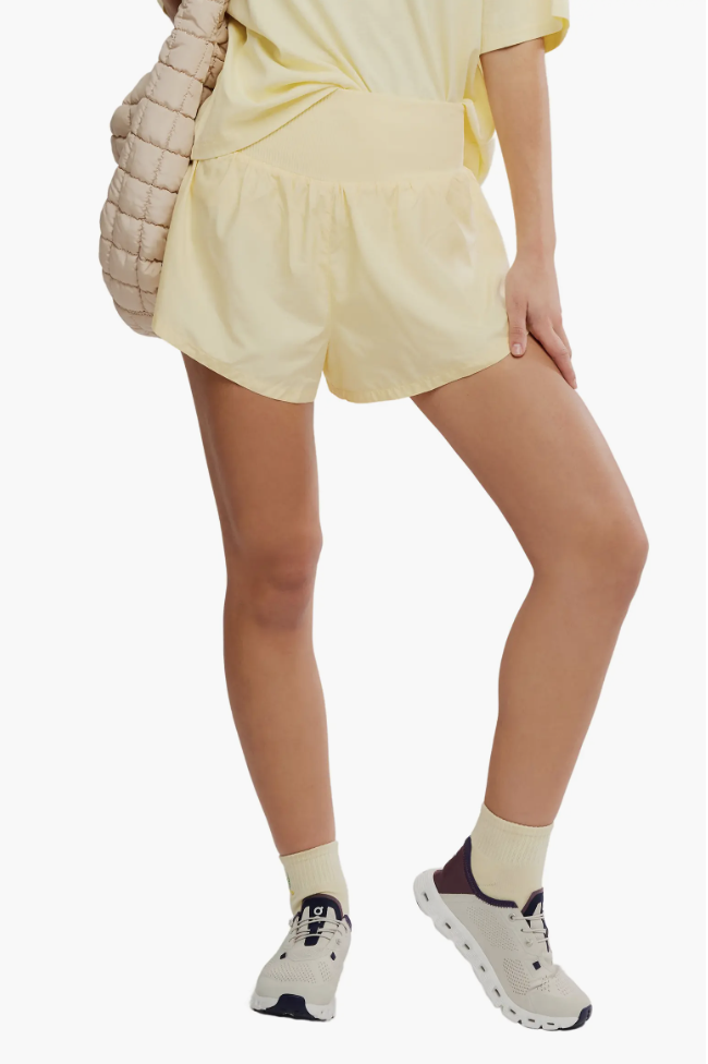 A close-up of a person wearing pastel yellow FP Movement Carpe Diem track shorts with a matching yellow top, holding a cream woven shoulder bag.