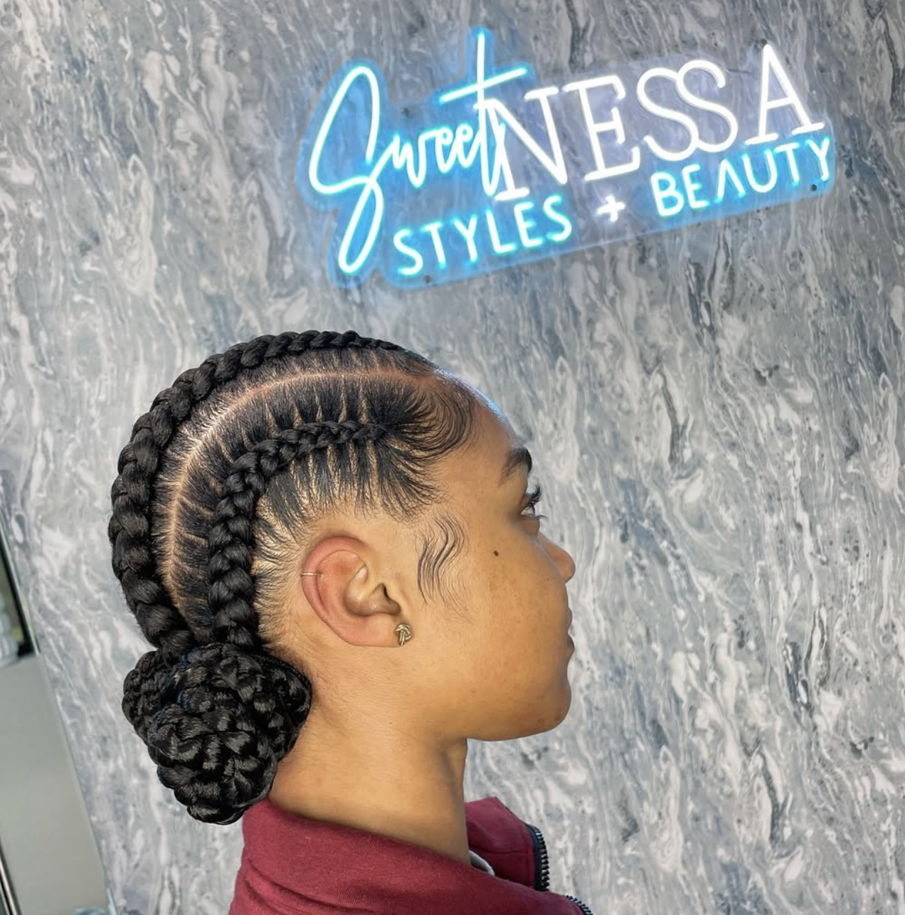 Side profile of a woman with neat, straight-back cornrows styled into a low braided bun, featuring detailed baby hairs and a gold earring.