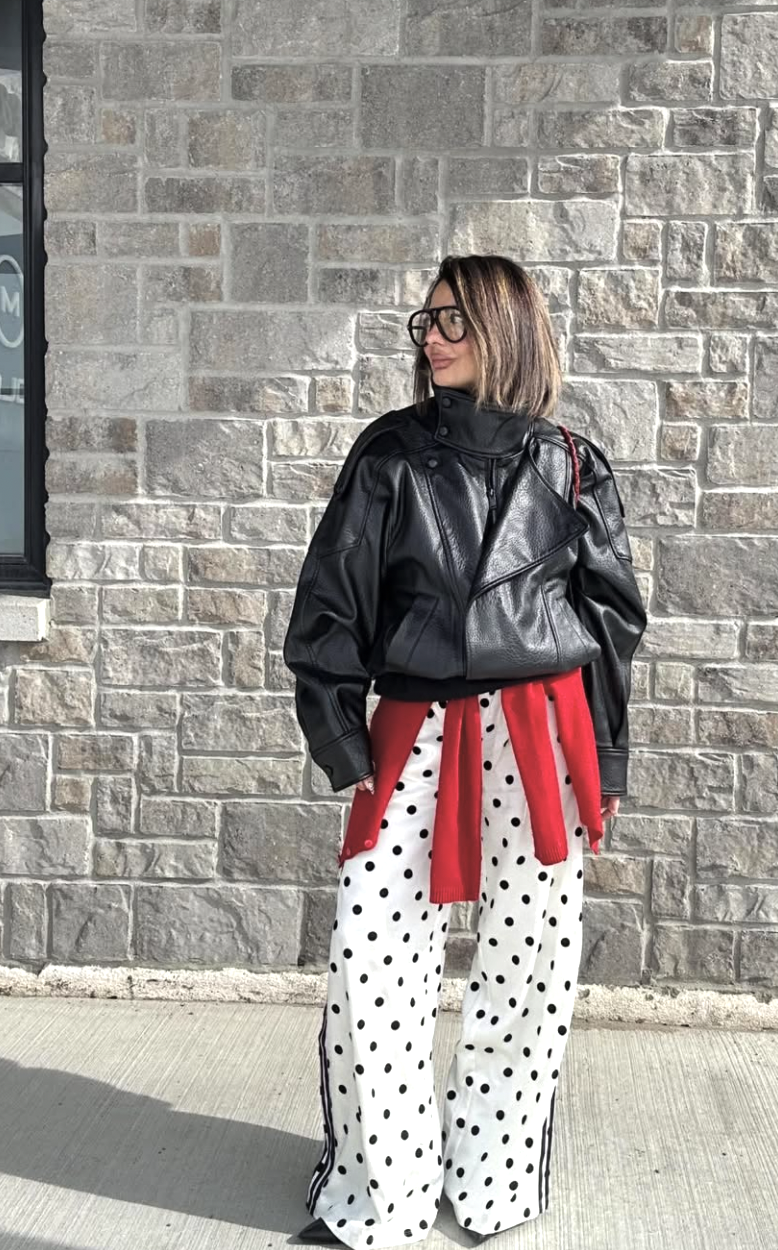 A woman wearing a black oversized leather moto jacket and white pants with large black polka dots, with a vibrant red sweater tied diagonally across her chest as a styling accent.