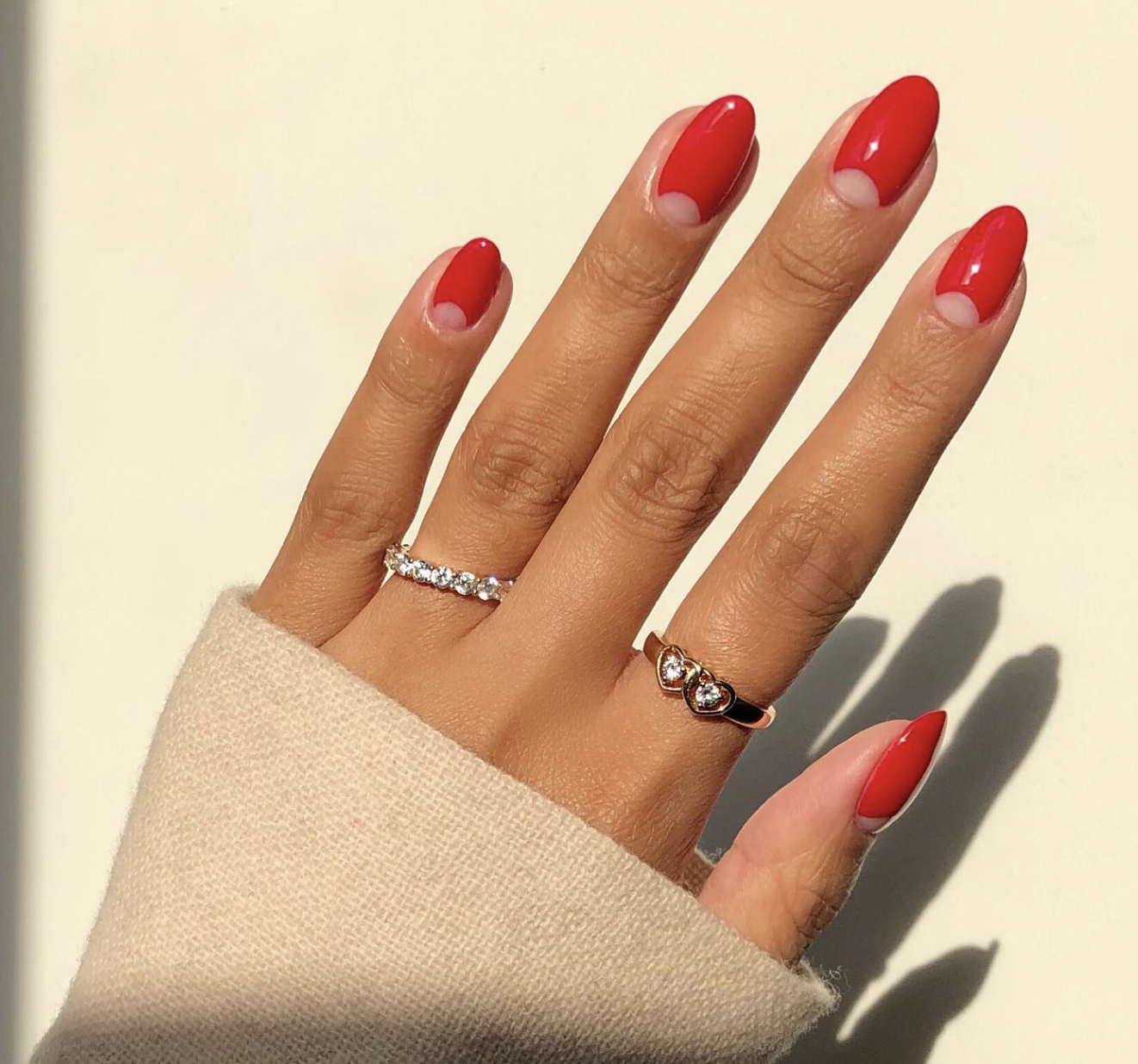 A close-up of a hand with a red half-moon manicure on a sheer base, featuring gold rings and a cream sweater.