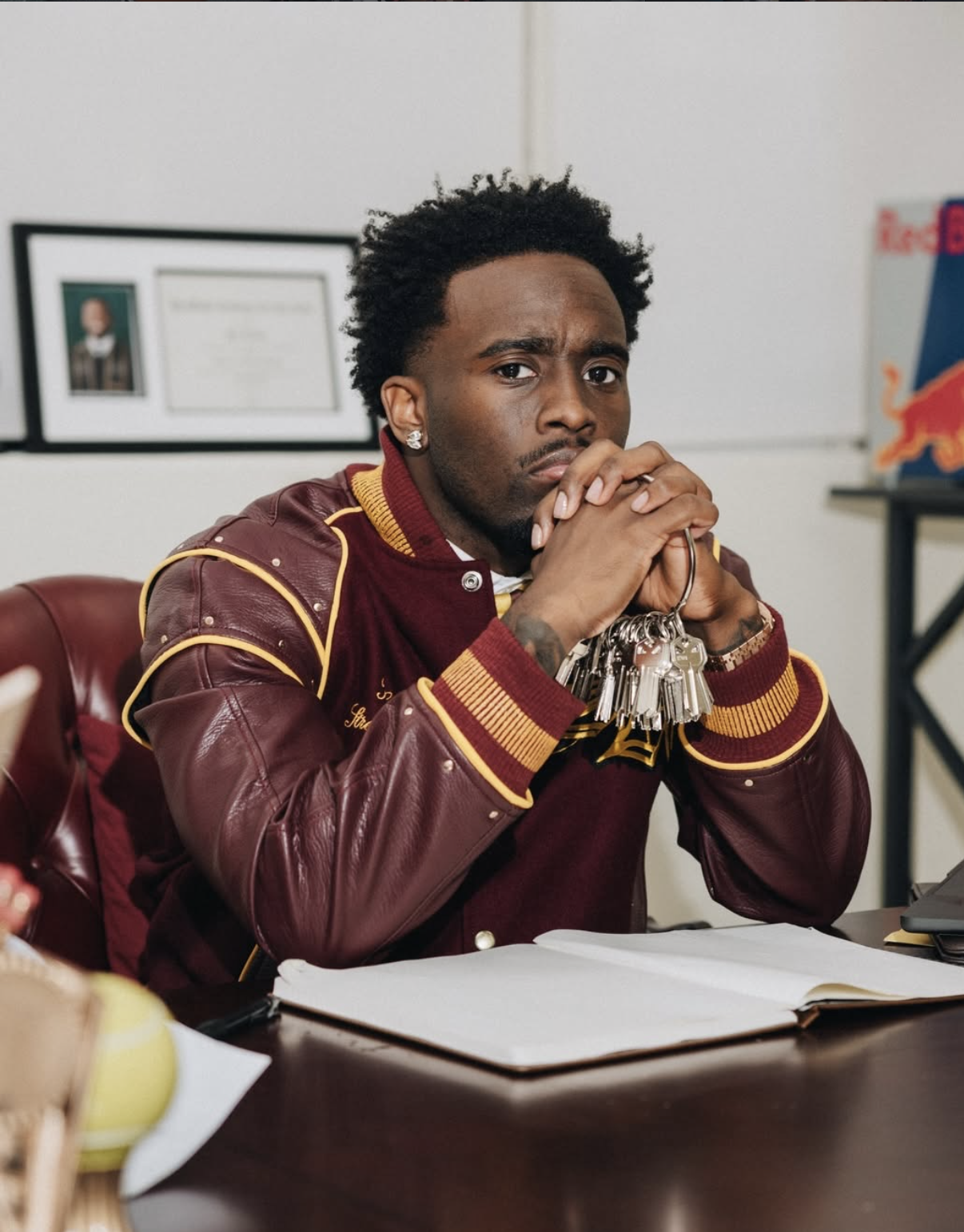 Kai Cenat sitting at a desk wearing a burgundy leather jacket with yellow trim.