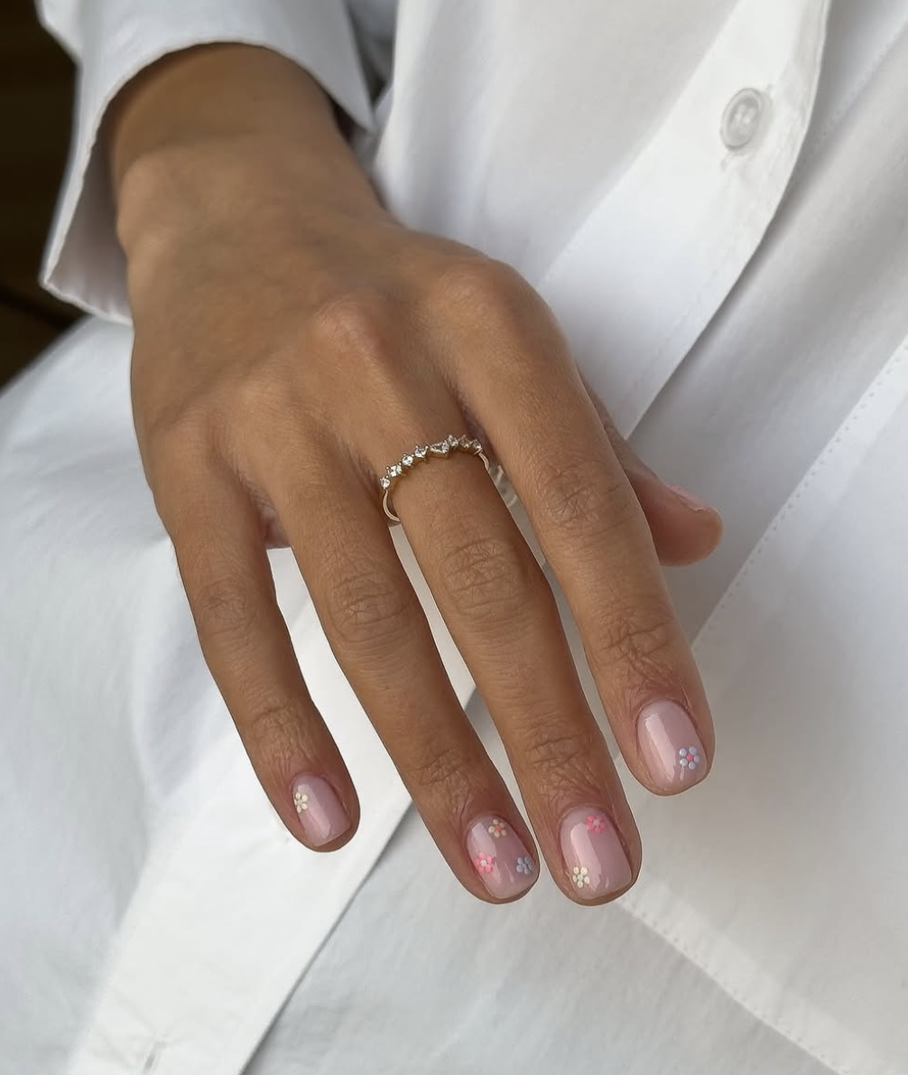 A close-up of a hand with short, natural-shaped nails featuring a high-shine sheer nude base and tiny, multi-colored hand-painted daisies in pastel blue, pink, and white.