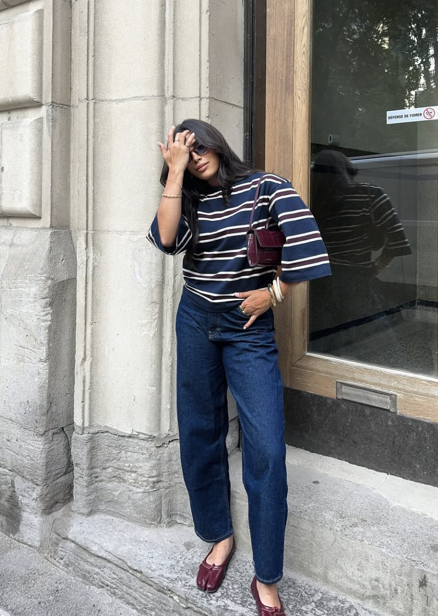 A person wearing dark wash barrel jeans paired with burgundy Tabi ballet flats, a striped navy and white sweater, and a matching burgundy shoulder bag.