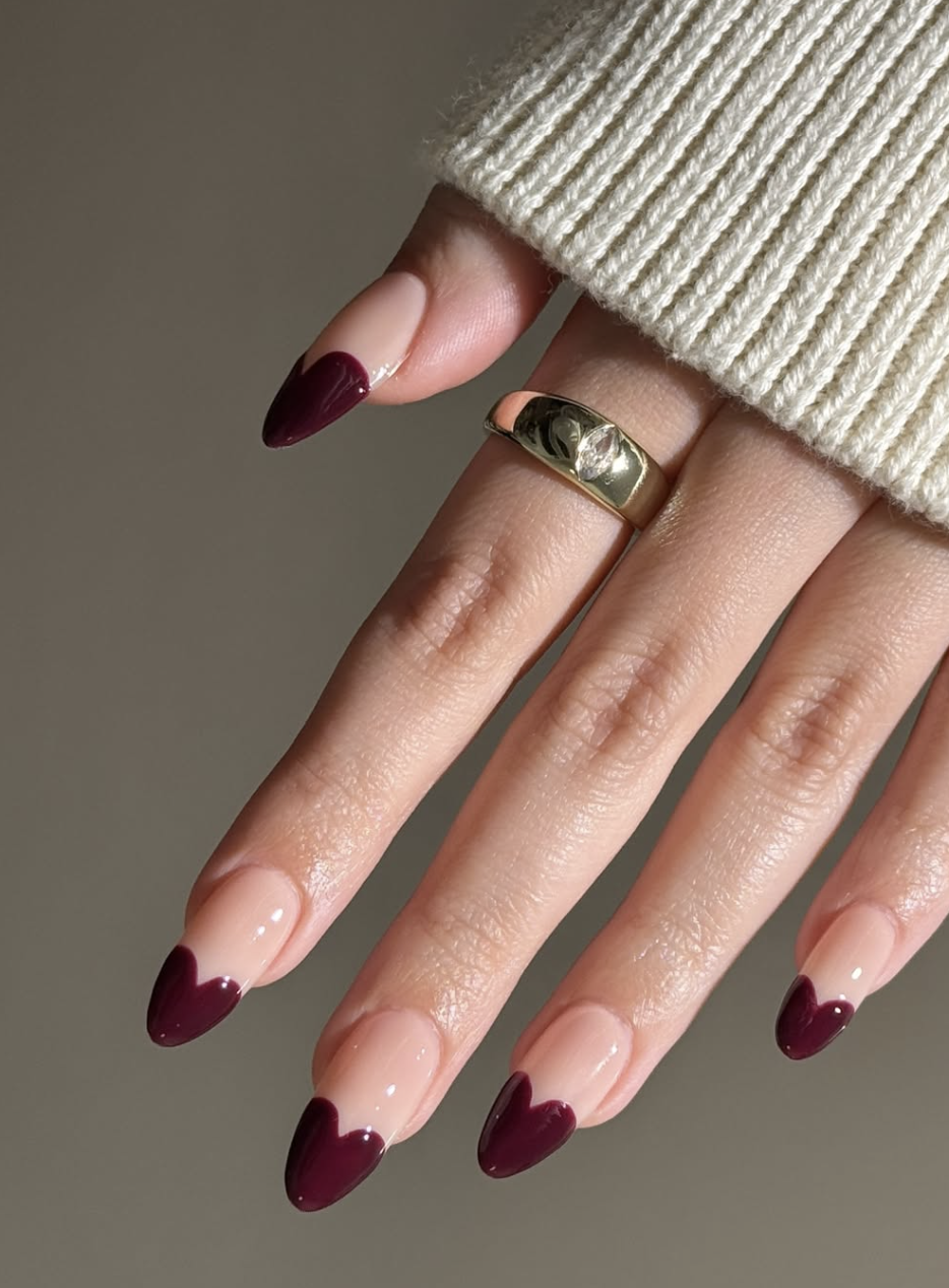 Almond-shaped nails with a sheer nude base and deep burgundy red heart-shaped French tips.