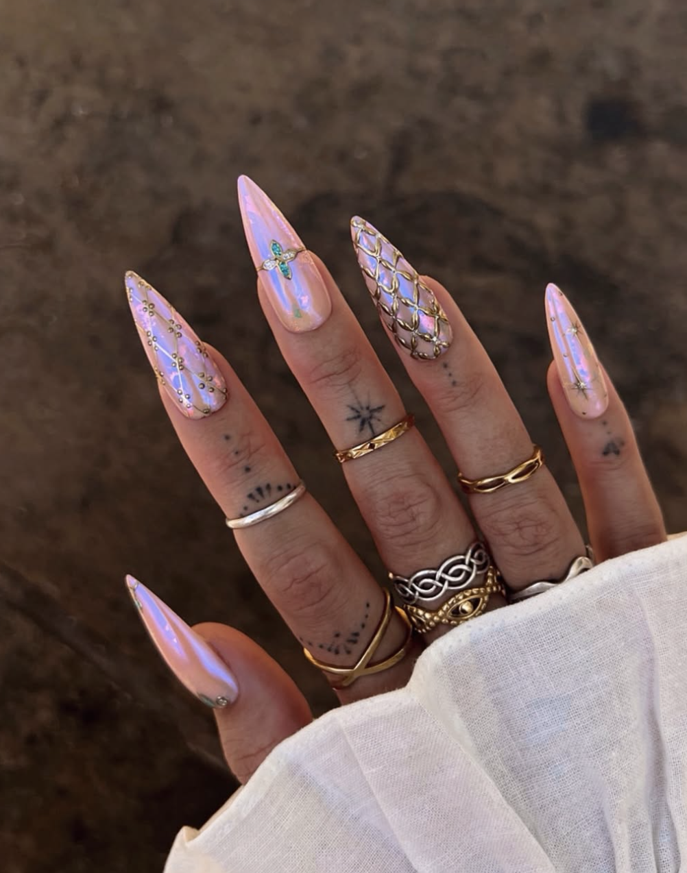 Close-up of a hand with long stiletto-shaped nails showcasing a futuristic iridescent pink manicure, featuring detailed 3D gold textured grid patterns, celestial star accents, and tiny turquoise center gems, accessorized with heavy gold rings.