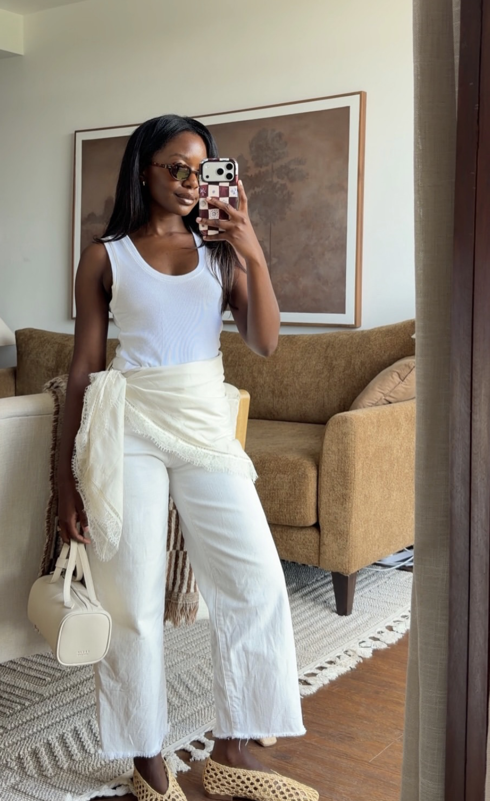 A woman styling a monochrome summer outfit featuring a white tank top, wide-leg linen trousers, a cream fringed wrap waist scarf, and neutral jelly flats.