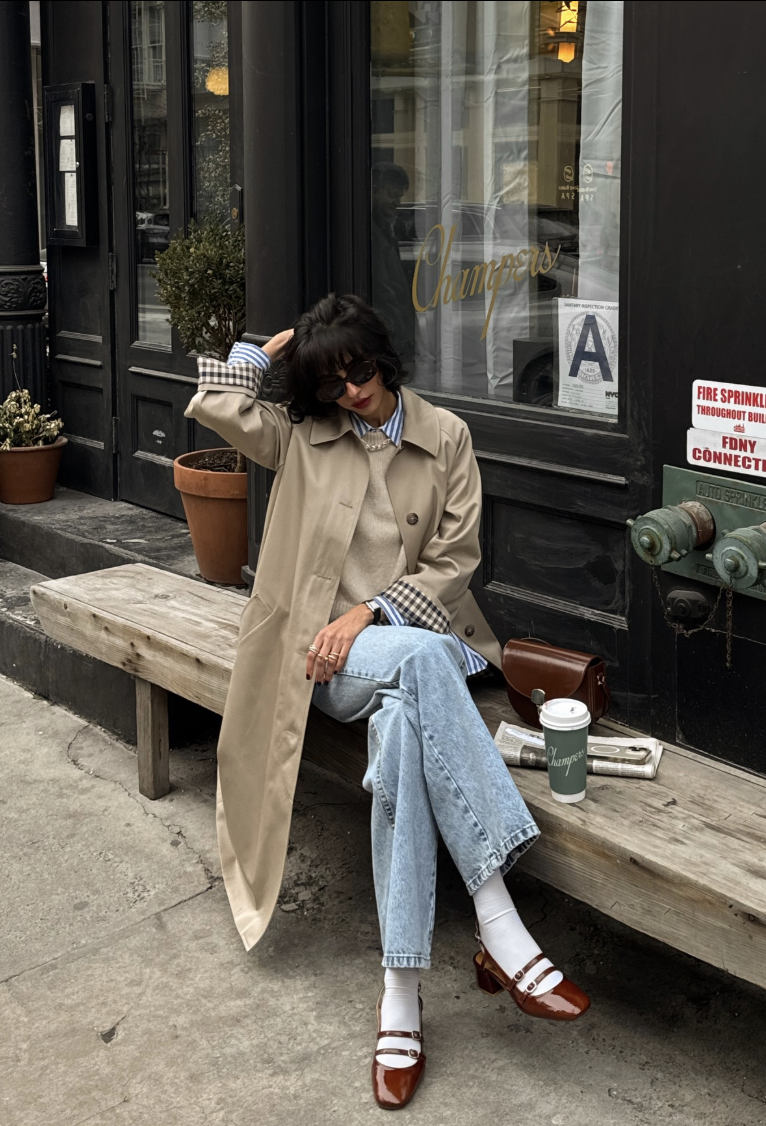 A woman wearing a beige trench coat over a striped shirt and sweater, paired with light wash jeans and white square-toe Mary Jane flats with a thin strap.