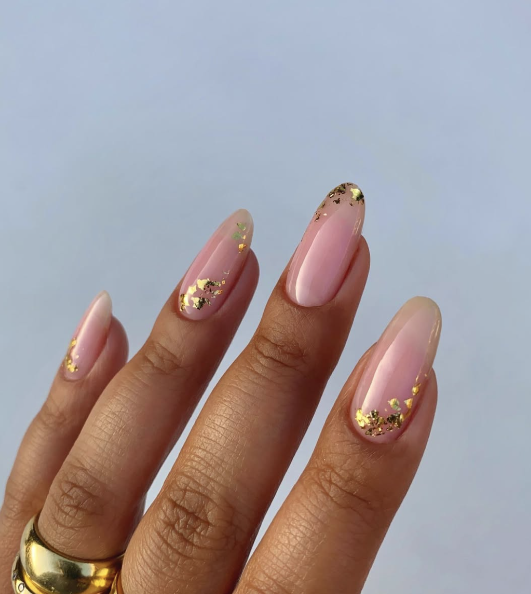 A close-up of long, oval-shaped nails with a sheer pink base, featuring a classic French manicure outline accented with shimmering flakes of gold foil, paired with a chunky gold ring.