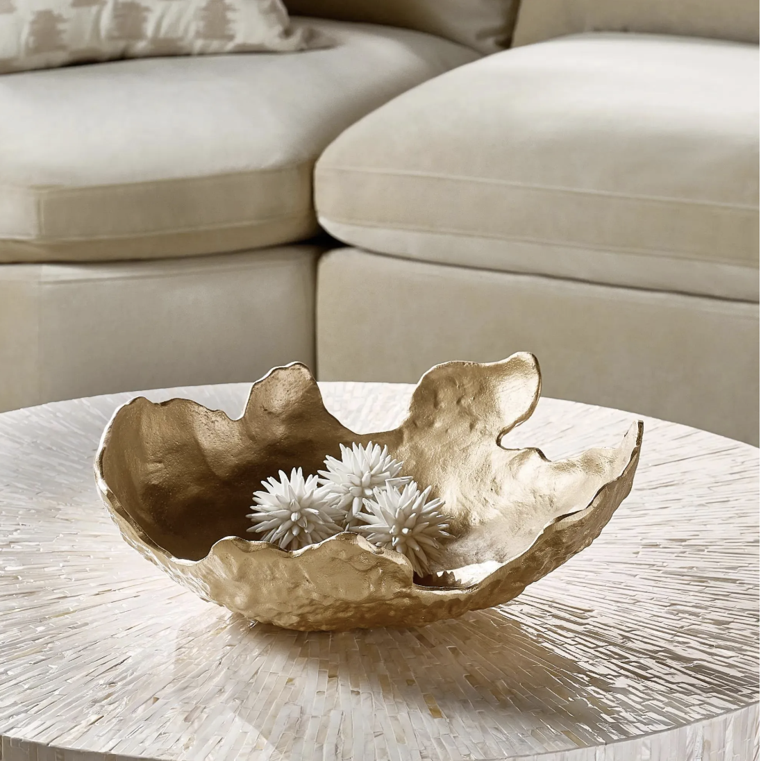 Crushed gold aluminum decorative bowl filled with white accents on a round coffee table in front of a cream sofa.