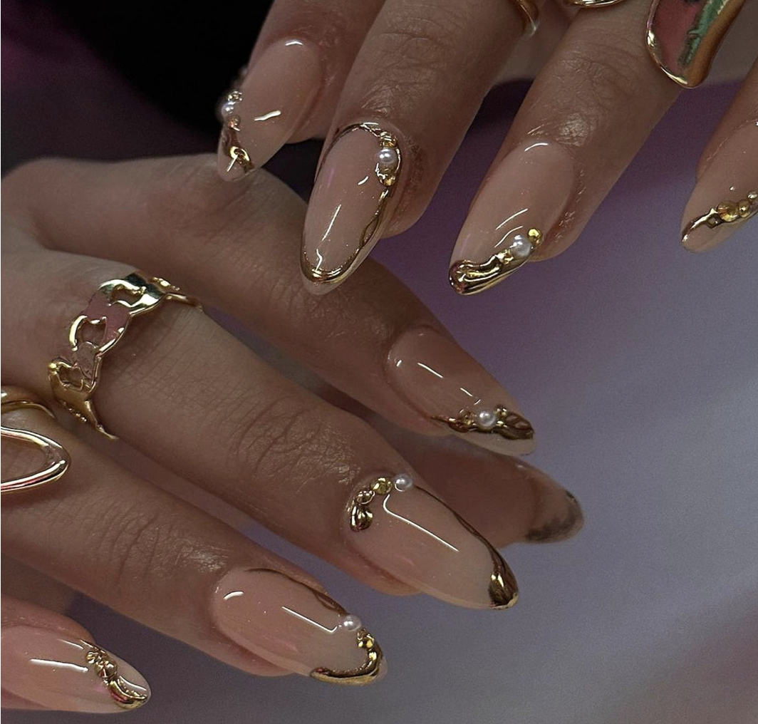 Medium almond-shaped press-on nails with a nude translucent base, featuring 3D gold chrome swirls and delicate white pearl accents.