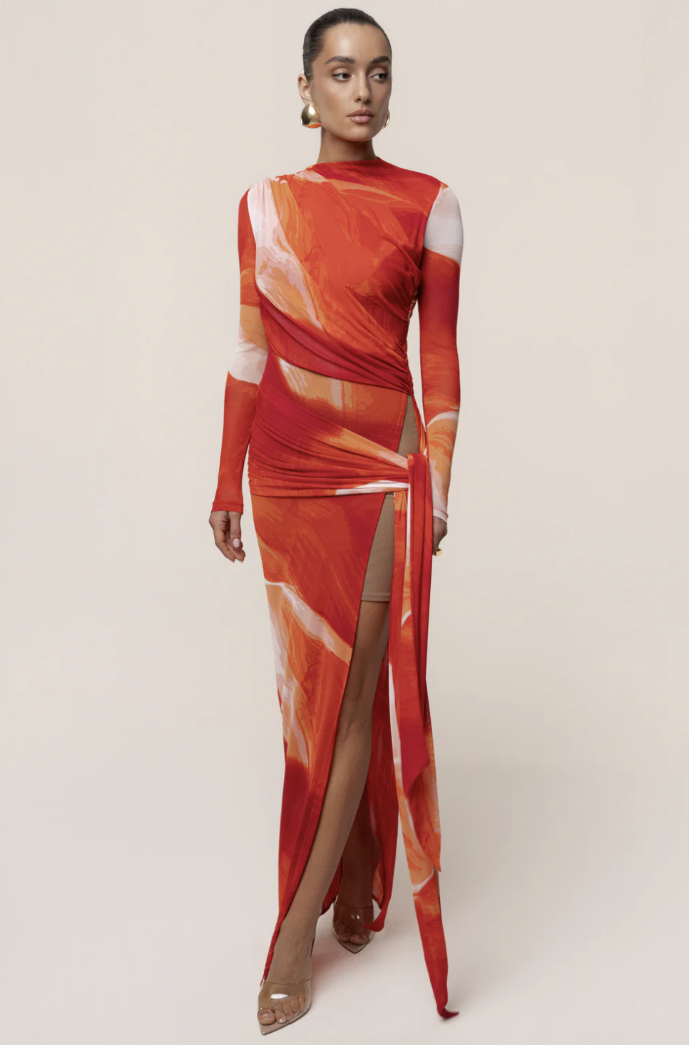 A woman wearing the JLUXLABEL Sunset Red Heatwave Mesh Maxi Dress with an abstract orange and red watercolor print and a high side slit, styled for a luxury resort vacation.