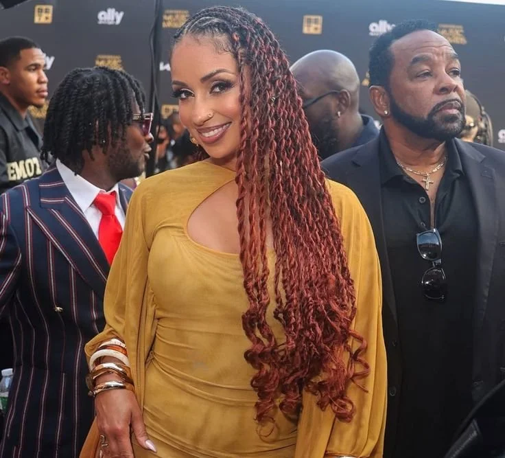 Mýa with long, copper-toned Senegalese twists falling over her shoulder, wearing a yellow dress and gold bangles to demonstrate how length creates a downward visual that balances a heart face shape.
