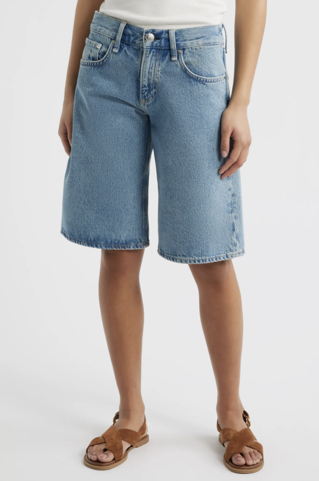 A woman wearing light-wash Gap 11-inch mid-rise longline denim Bermuda shorts with a raw hem, styled with brown crossover sandals.