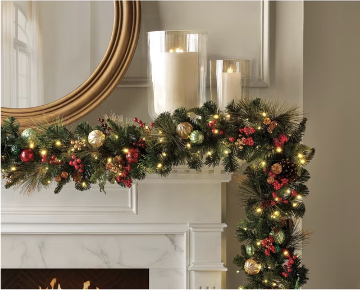 White fireplace mantel draped in a pre-lit evergreen garland with red berries and ornaments under a round gold mirror.