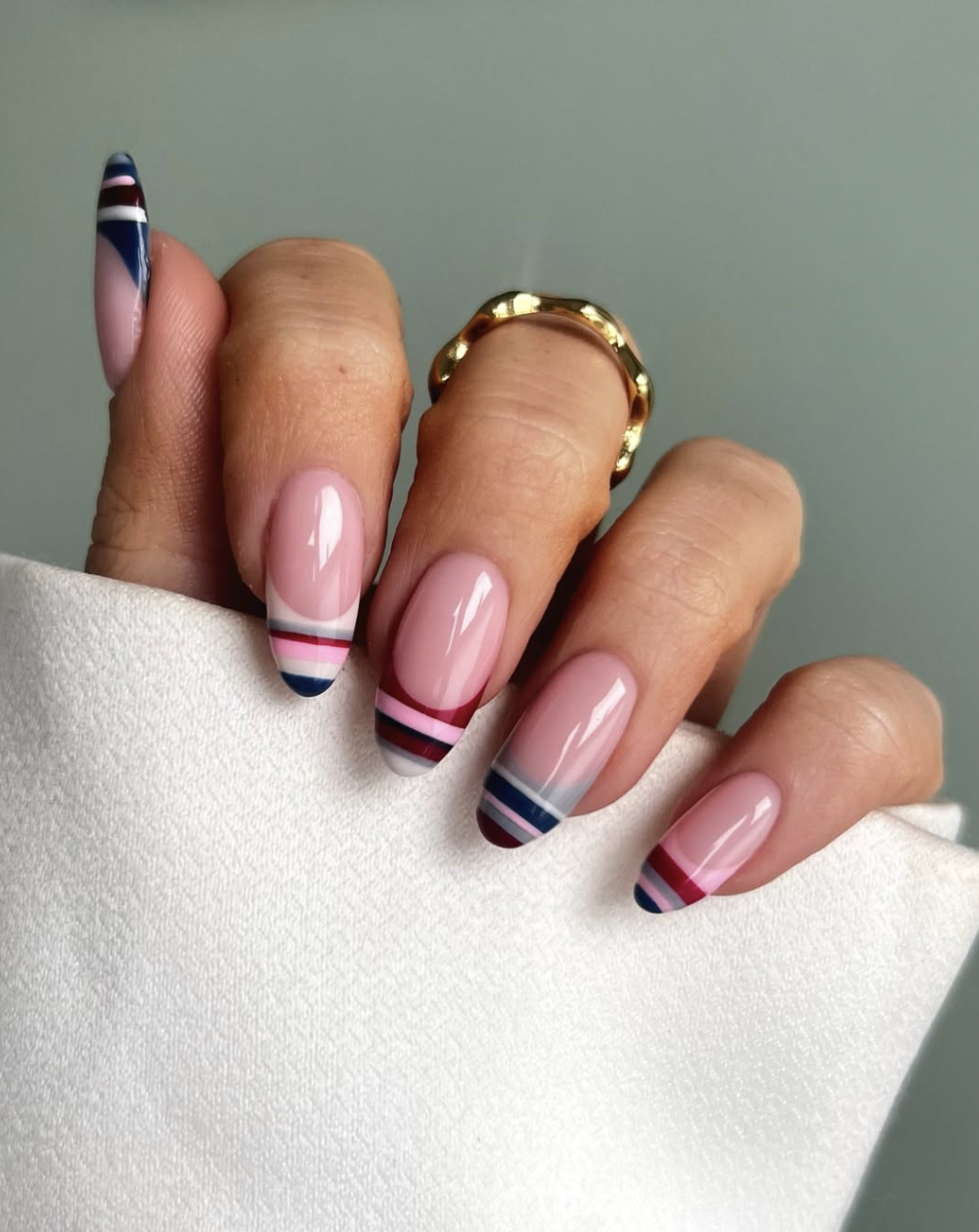 Long almond-shaped nails with a sheer nude base and structured, geometric French tips featuring stacked horizontal stripes in navy blue, burgundy, and soft blue.