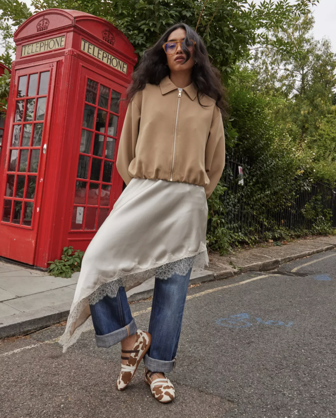 Woman standing on a London street next to a red telephone booth wearing a tan cropped bomber jacket over a white lace-trimmed asymmetric camisole and cuffed wide-leg jeans with animal print sandals.
