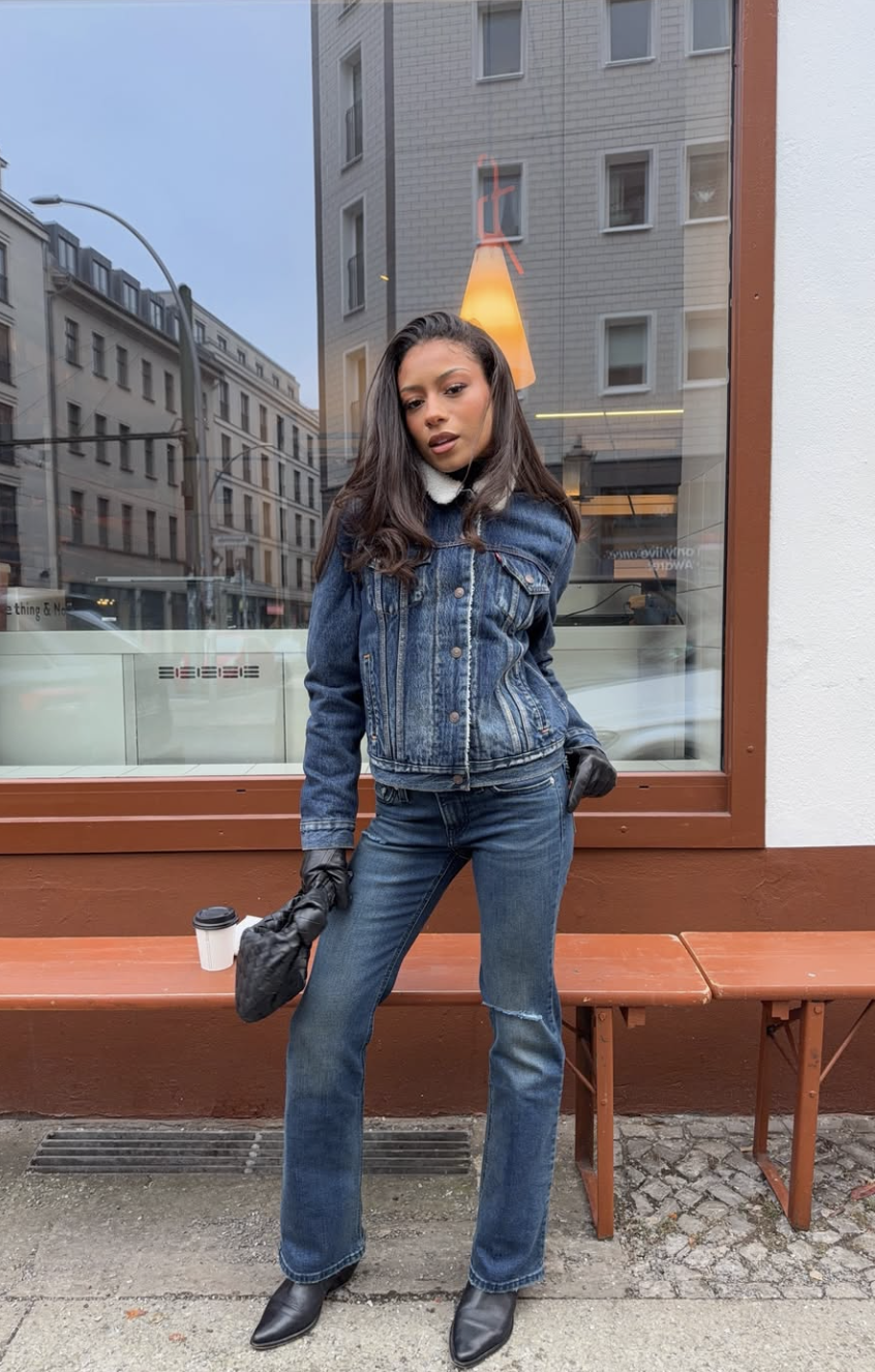 Woman wearing a matching denim shirt jacket and dark wash bootcut jeans with black pointed-toe boots and a black clutch.