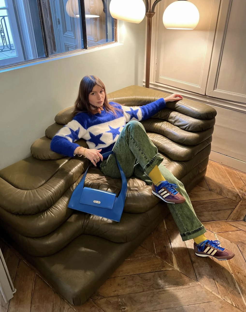 A woman sitting on an olive green leather sofa wearing a blue fuzzy sweater with large white stars, green trousers, bright yellow crew socks, and blue retro sneakers, holding a structured blue shoulder bag.