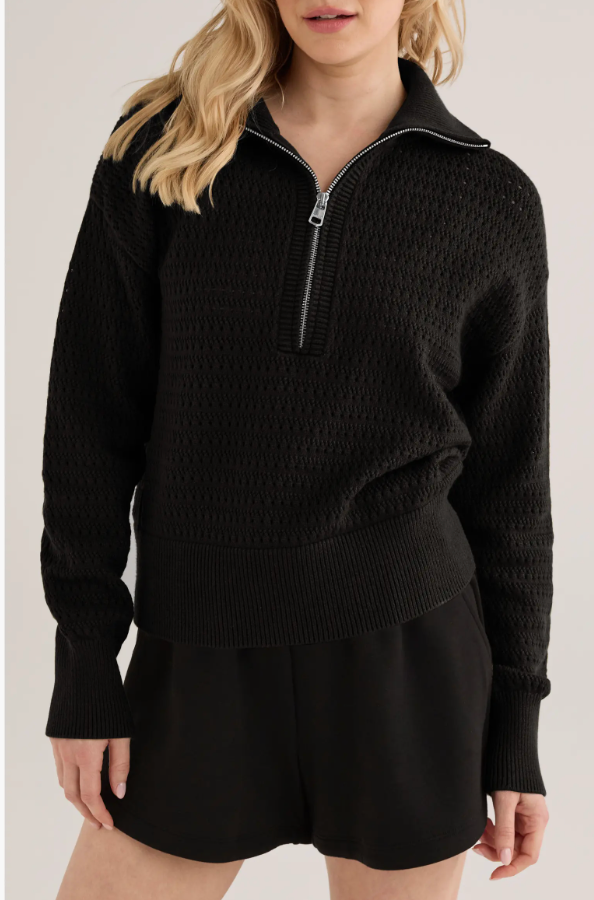 A close-up shot of a model wearing the Varley Darla Half-Zip Sweater in black, featuring a heavy knit texture and a silver zipper pulls, paired with matching black shorts.