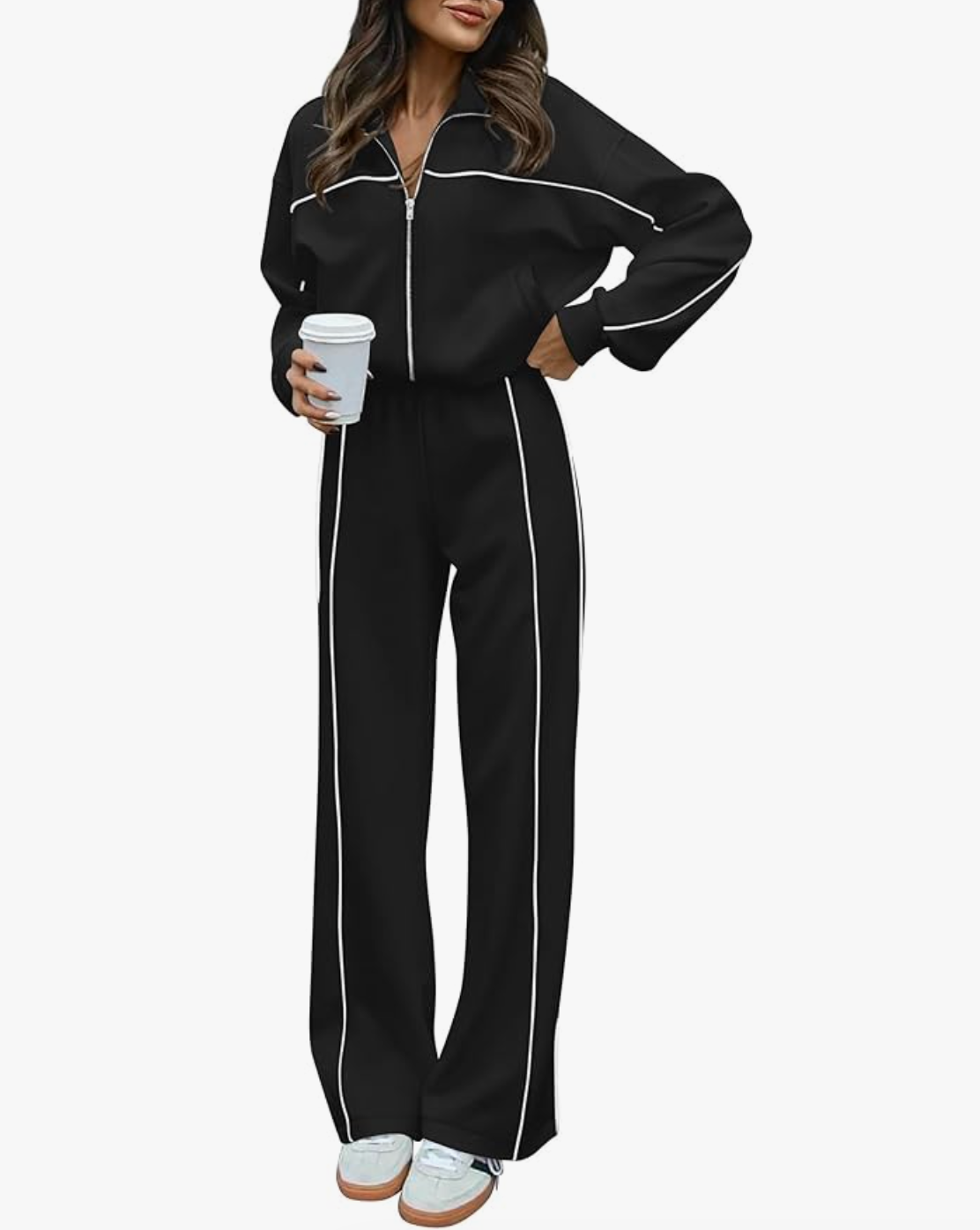 A woman wearing a black two-piece tracksuit with white piping details on the sleeves and legs, holding a white coffee cup, paired with white and grey sneakers.