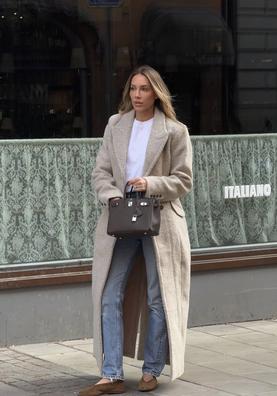 A street style lookbook image of a woman wearing a white t-shirt tucked into blue straight-leg jeans, layered under a long, textured oatmeal-colored wool coat.